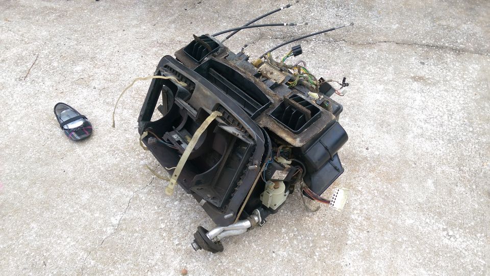 Disassembled car HVAC unit with wires and ducting on concrete with a child's shoe.