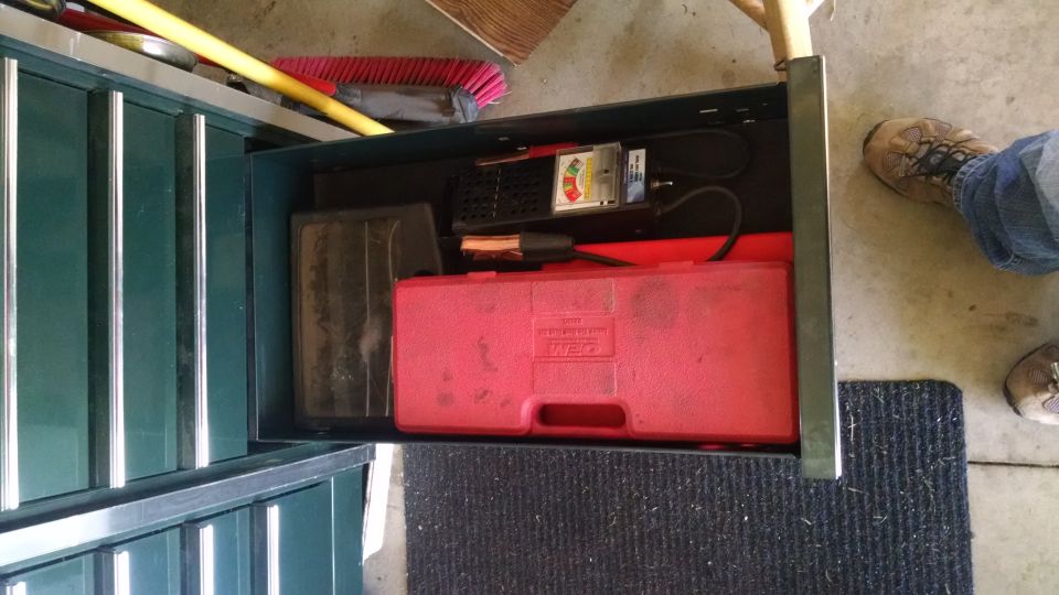 Open toolbox drawer with red tool case and battery tester, person's feet visible.