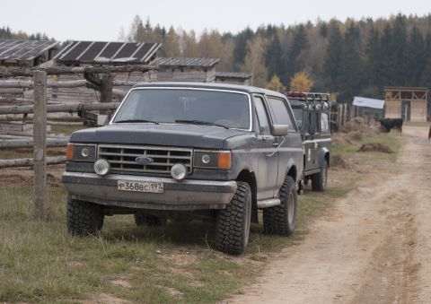 1987 Ford Bronco - A dark-colored Ford Bronco is parked on a dirt road next to a wooden fence and rustic buildings. Another off-road vehicle is parked behind…