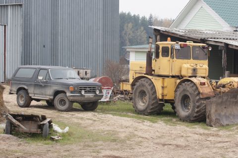 1987 Ford Bronco - A dark-colored, lifted Ford Bronco is parked next to a large yellow tractor with a front loader. The scene appears to be a rural or farm se…