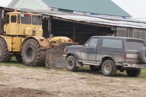 1987 Ford Bronco - A black Ford Bronco is parked next to a yellow tractor with a front loader. The vehicles are on a dirt and grass area in front of a rustic…