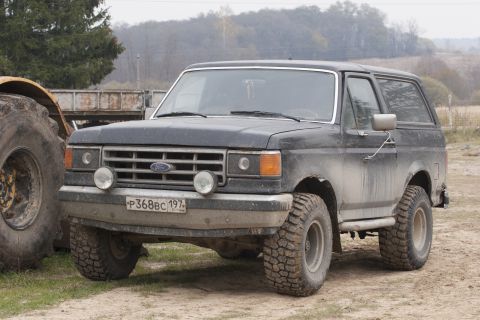 1987 Ford Bronco - A dark-colored, lifted Ford Bronco sits parked on a dirt road next to a large tractor tire. The vehicle has off-road tires and a license pl…