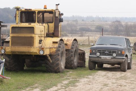 1987 Ford Bronco - A yellow articulated tractor and a dark-colored Ford Bronco are parked side-by-side on a dirt and grass area with a rural landscape in the…