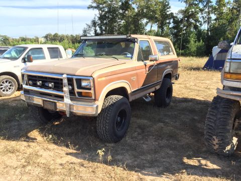 1996 Ford Bronco - A brown two-door Ford Bronco with a chrome brush guard and a light bar mounted on the roof is parked on a dirt lot next to a white pickup t…