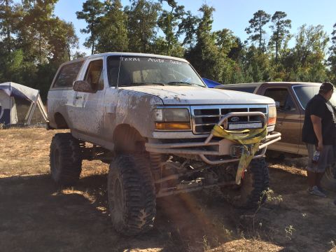 1996 Ford Bronco - A white, mud-covered 1990s Ford Bronco is parked outdoors with a tent and trees in the background. The vehicle has large off-road tires, a…