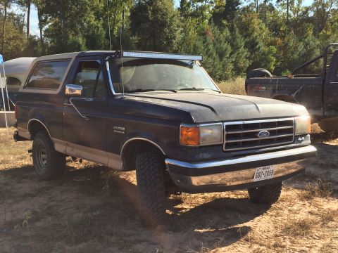 1996 Ford Bronco - A dark blue Ford Bronco is parked on a dirt lot with trees in the background. The vehicle has a tan lower body trim, a light bar mounted on…