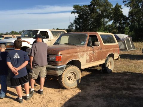 1996 Ford Bronco - A muddy, brown, two-door 1996 Ford Bronco is parked on a dirt lot next to a white Ford truck. A tent is set up in the background.