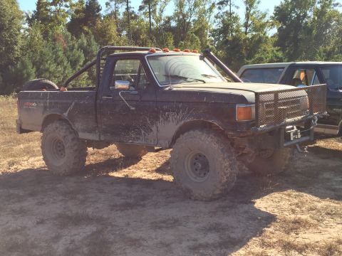 1996 Ford Bronco - A muddy, black, lifted Ford pickup truck with large off-road tires and a roll cage is parked on a dirt path with trees in the background. A…