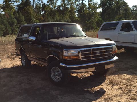 1996 Ford Bronco - A black 1996 Ford Bronco is parked outdoors on a dirt lot with trees in the background. A white Ford Bronco is parked behind it.