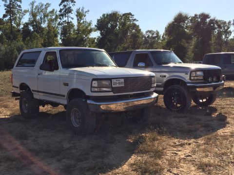1996 Ford Bronco - A lifted white 1996 Ford Bronco is parked on a dirt lot next to a silver Ford Bronco. The white Bronco has chrome trim and aftermarket whee…