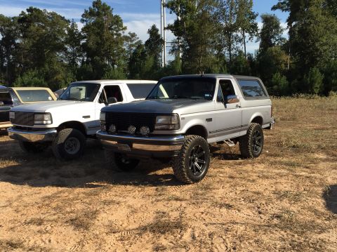 1996 Ford Bronco - Two Ford Broncos are parked on a dirt lot with trees in the background. The Bronco in the foreground is silver and has aftermarket wheels a…