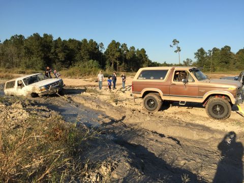 1996 Ford Bronco - Two Ford Broncos are stuck in mud on a sunny day. The white Bronco is partially submerged, while the brown Bronco is being pulled out by a…
