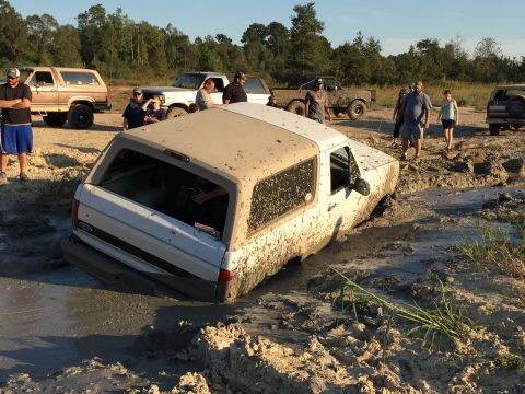 1996 Ford Bronco - A white Ford Bronco is stuck in deep mud with several people standing around watching. Other vehicles and trees are visible in the backgrou…