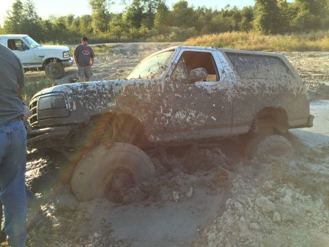 1996 Ford Bronco - A heavily mud-covered Ford Bronco is stuck in deep mud with its front tires submerged. Another white pickup truck is visible in the backgro…