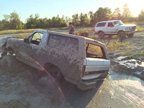 1996 Ford Bronco - A heavily mud-covered Ford Bronco is stuck in thick mud with a white Ford Bronco visible in the background.