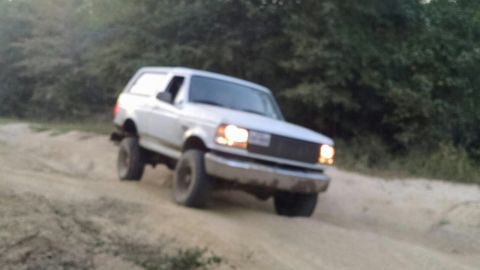 1996 Ford Bronco - A white 1990s Ford Bronco is driving off-road on a dirt path. The vehicle is angled towards the right side of the frame with its headlights…