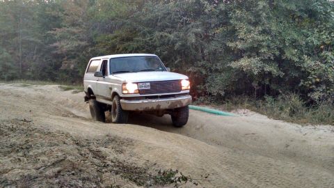 1996 Ford Bronco - A white 1990s Ford Bronco is driving on a sandy, off-road trail surrounded by trees. The Bronco's headlights are on, and it is angled towar…