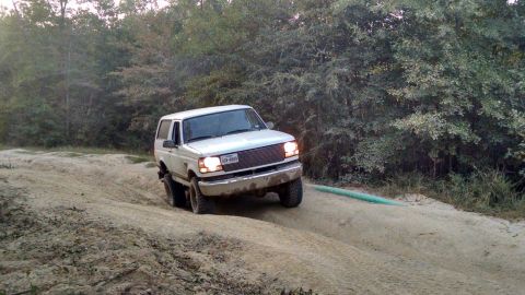 1996 Ford Bronco - A white 1996 Ford Bronco is driving on a sandy, off-road trail with trees in the background. The vehicle is angled towards the camera, show…