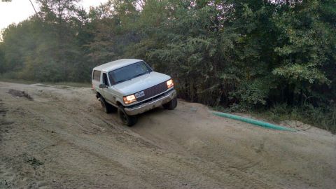 1996 Ford Bronco - A white 1996 Ford Bronco is driving up a sandy incline on a dirt road, surrounded by trees.