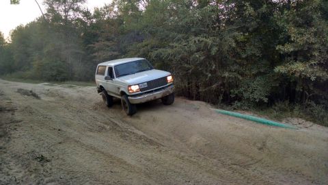 1996 Ford Bronco - A white 1990s Ford Bronco is driving up a sandy incline next to a line of trees. The vehicle is angled towards the viewer and its headlight…