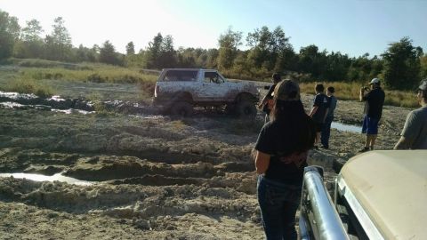 1996 Ford Bronco - A lifted, mud-covered white Ford Bronco is stuck in a muddy track with several people watching. The scene appears to be an off-road park or…