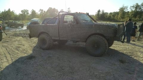 1996 Ford Bronco - A heavily mud-covered pickup truck is parked on a dirt track with trees in the background. Several people are standing around the truck.