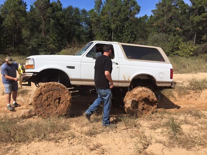 White Ford Bronco stuck in mud with large tires, two men nearby.