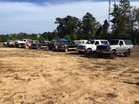 1996 Ford Bronco - A line of Ford Bronco SUVs and pickup trucks are parked on a dirt lot with trees and a water tower in the background.