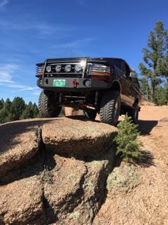 1996 Ford Bronco - A black 1996 Ford Bronco is shown from a low angle, driving on a dirt road over large rocks with a blue sky and trees in the background. Th…