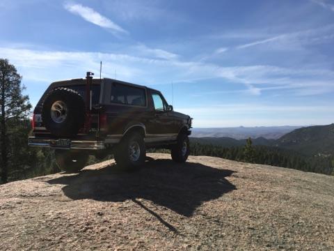 1996 Ford Bronco - A dark red 1996 Ford Bronco is parked on a rocky outcrop overlooking a vast, forested landscape under a bright blue sky with scattered clou…