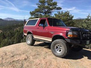 1996 Ford Bronco - A red 1996 Ford Bronco is parked on a dirt hill with a mountainous, wooded landscape in the background under a clear blue sky.