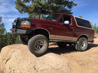 1996 Ford Bronco - A maroon 1996 Ford Bronco with a tan top is parked on a large rock formation, angled towards the viewer's left. The SUV features a brush gu…