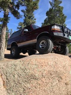 1996 Ford Bronco - A maroon 1996 Ford Bronco is positioned on a large rock formation with pine trees and a blue sky in the background. The vehicle is viewed f…