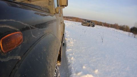 1987 Ford Bronco - A dark colored SUV drives through a snowy field, with another SUV visible in the distance.