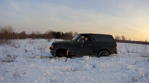 1987 Ford Bronco - A dark colored SUV is stuck in deep snow in a field with trees in the background. A person is visible driving the vehicle.