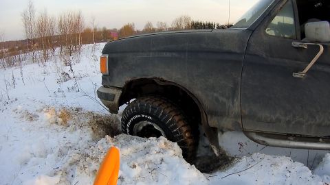 1987 Ford Bronco - A dark-colored SUV is stuck in deep snow on a snowy embankment. The front driver's side tire and fender are visible, along with part of the…