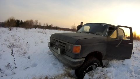 1987 Ford Bronco - A dark-colored pickup truck is stuck in the snow with its driver's side door open. The truck is facing the camera at a three-quarter front…
