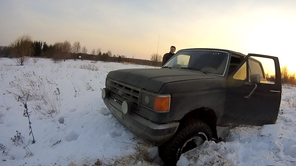Black pickup truck stuck in snow, driver's side door open, sunset sky.