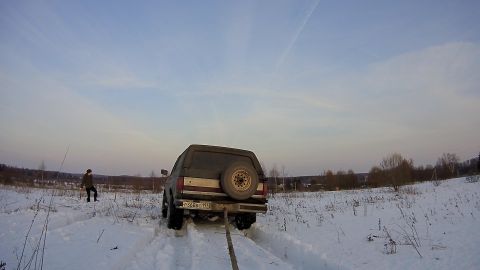 1987 Ford Bronco - The rear of a dark-colored SUV is stuck in the snow, with a tow rope attached to its bumper. A person stands in the snowy field to the left…