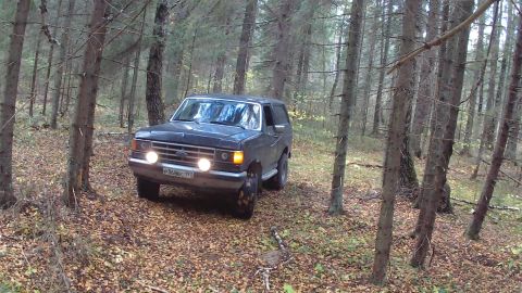1987 Ford Bronco - A dark-colored Ford Bronco is driven off-road through a forest with fallen leaves on the ground. The vehicle's headlights and turn signals…