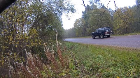 1987 Ford Bronco - A dark-colored SUV with a spare tire mounted on the rear is driving on a wet asphalt road bordered by trees and grass.
