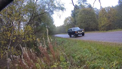 1987 Ford Bronco - A dark colored SUV drives down a paved road bordered by trees with fall foliage. The vehicle is viewed from the front-right quarter.