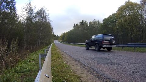 1987 Ford Bronco - A black Ford Bronco is driving down a paved road lined with trees and a guardrail. The vehicle has a spare tire mounted on the rear.