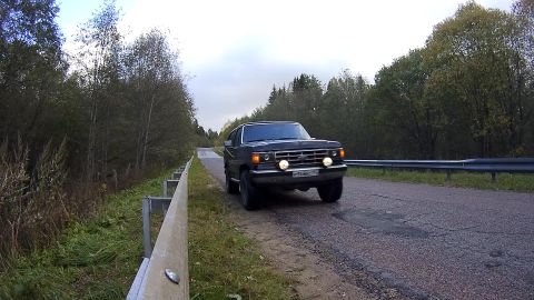 1987 Ford Bronco - A dark-colored Ford Bronco is driving on a paved road lined with trees and a guardrail. The vehicle is angled towards the camera, showing i…