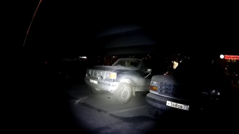 1987 Ford Bronco - Two dark-colored SUVs are parked at night. The SUV in the foreground on the left has its headlights on, illuminating the front end and a li…