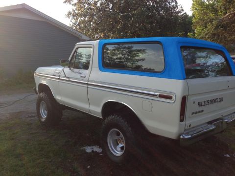 1994 Ford Bronco - The rear three-quarter view of a lifted, white Ford Bronco with a blue soft top is parked on a grassy area next to a building. The text "FU…