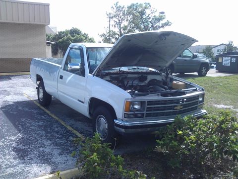 1994 Ford Bronco - A white pickup truck with its hood open is parked in a lot next to a building. The truck's engine is visible, and there is a green SUV park…