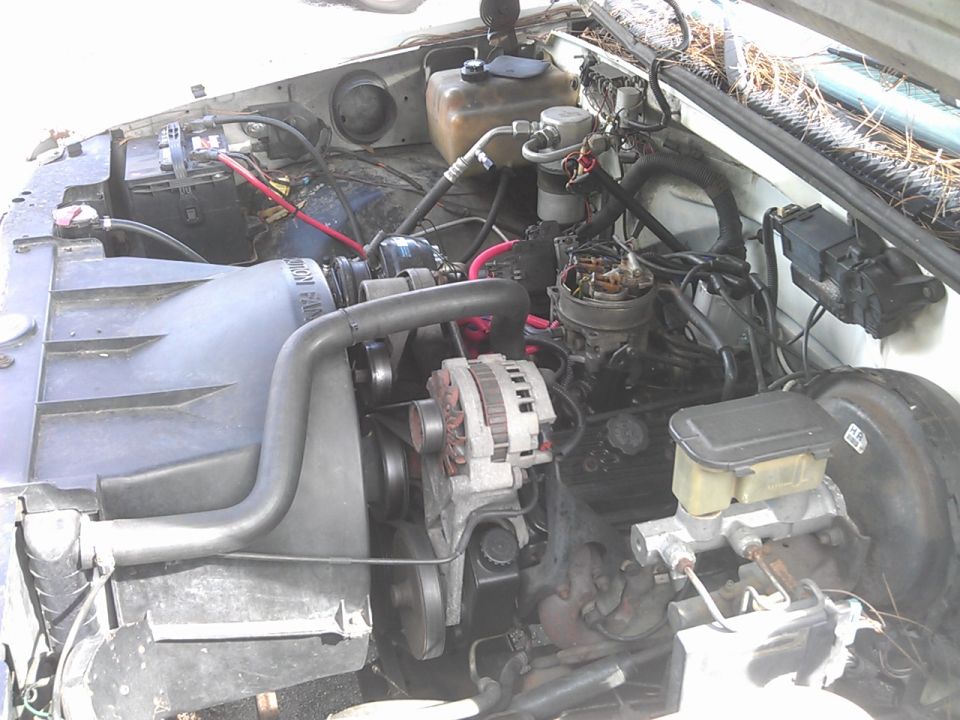 Engine bay of a vehicle with radiator fan, alternator, carburetor, and battery visible.