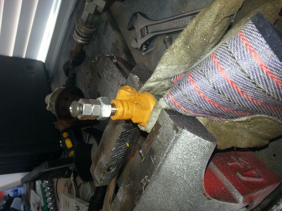 Close-up of an engine part with a yellow connector and metal fittings, held in a vise