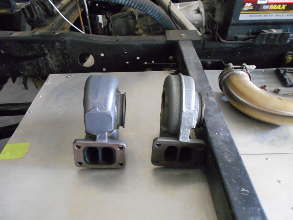 Two turbochargers and exhaust piping on a metal surface with vehicle frame in background